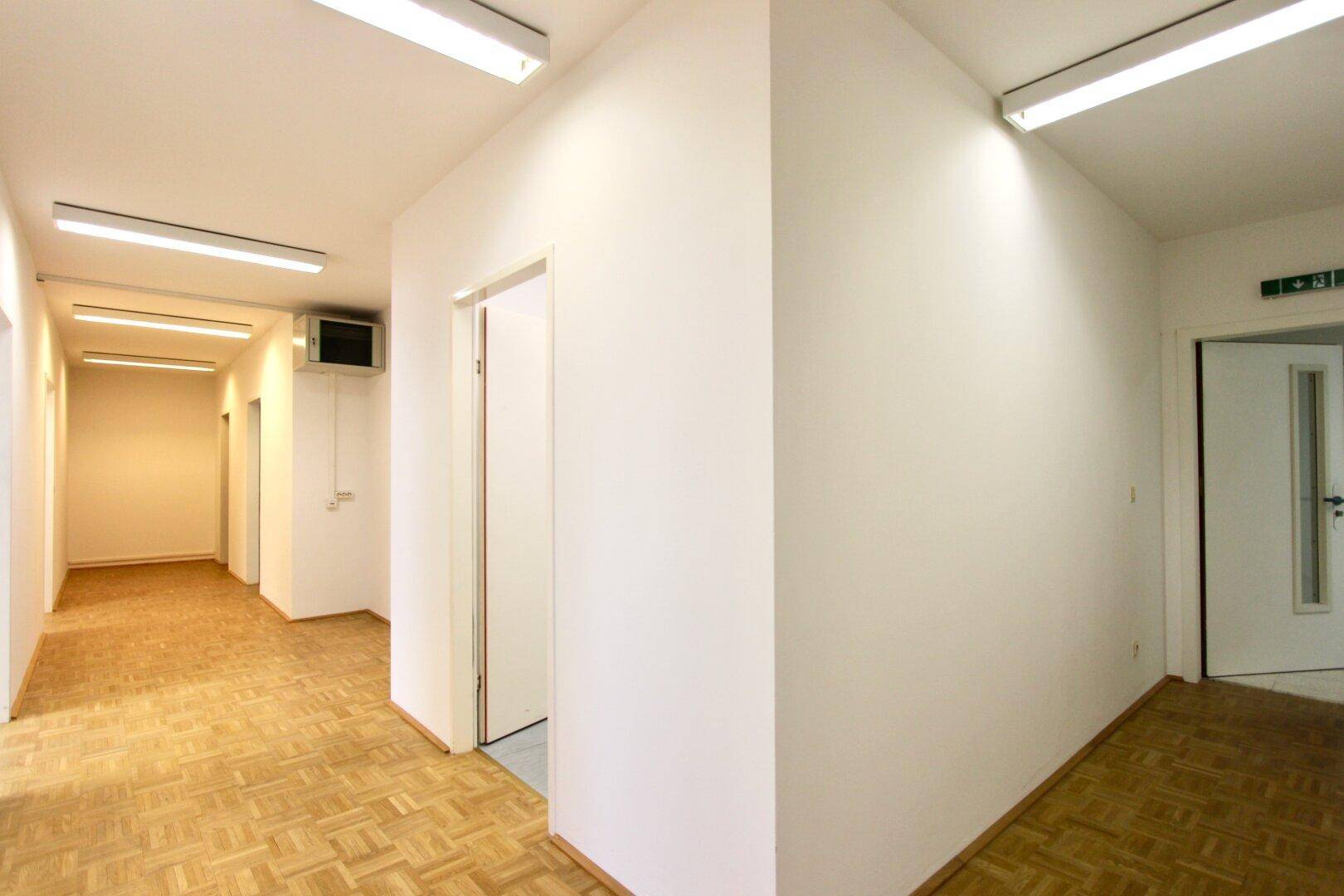 Langer, heller Flur mit Parkettboden und mehreren Türen, beleuchtet von Deckenleuchten.
