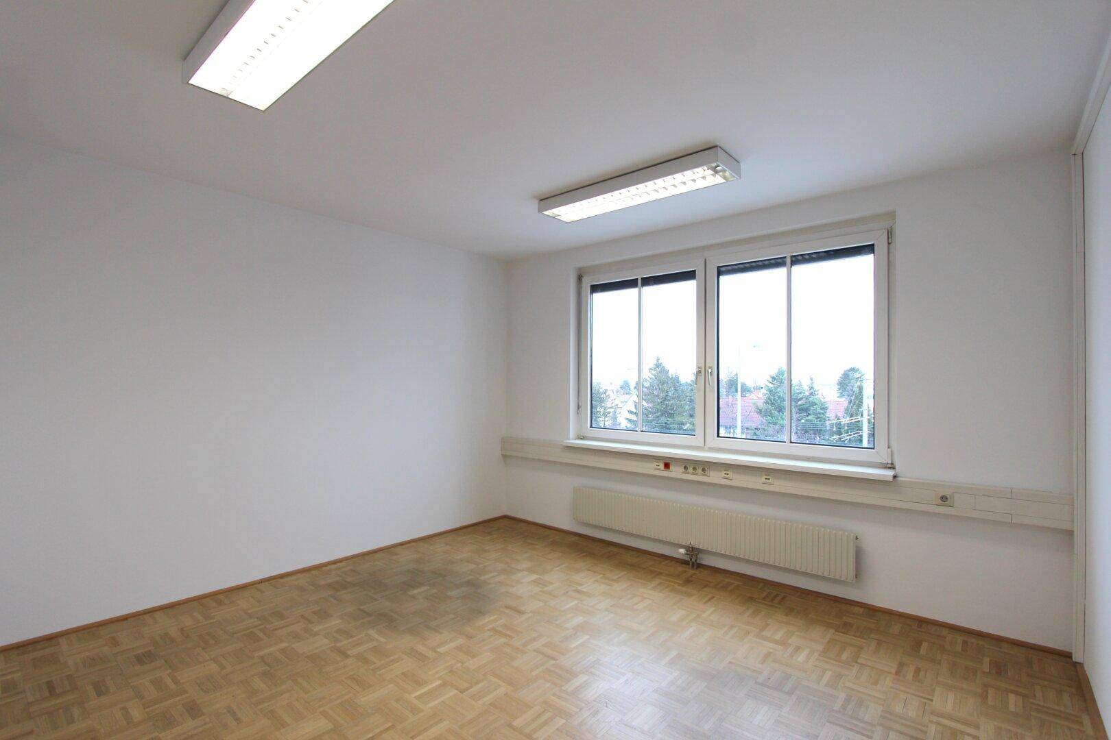 Leeres Büro mit Parkettboden, großem Fenster und Heizkörper unter der Fensterbank.