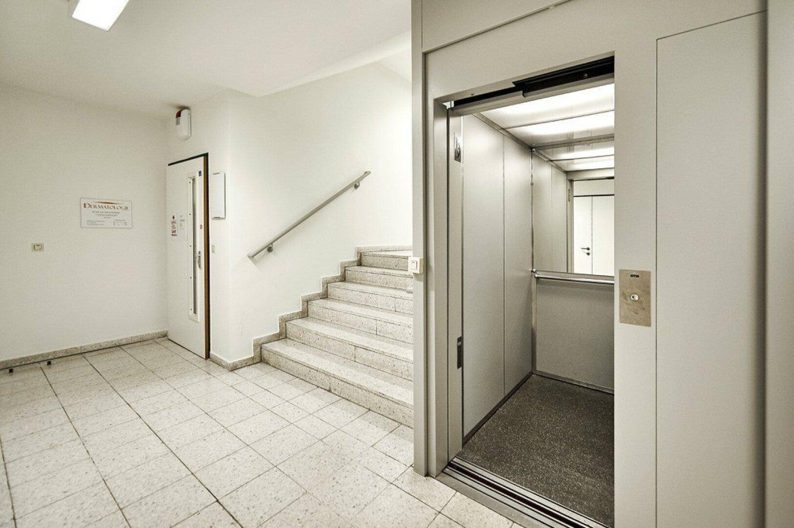 Moderner Eingangsbereich mit offenem Aufzug und Treppe, der eine barrierefreie Erreichbarkeit gewährleistet.