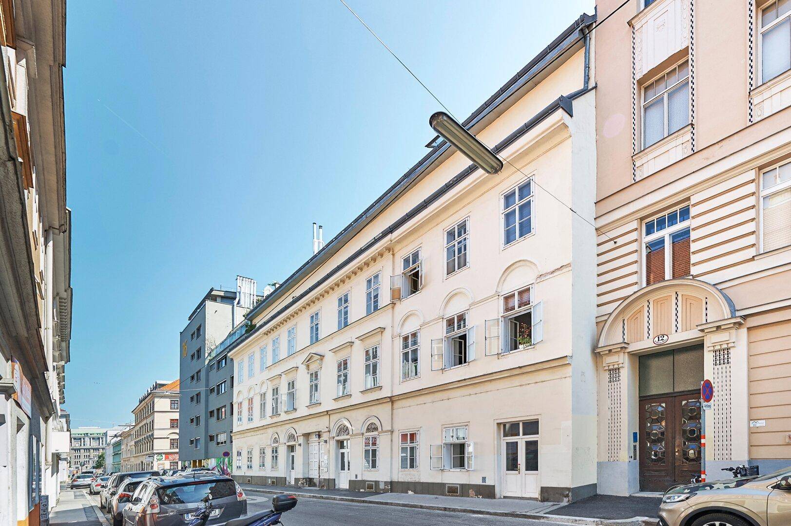 Lange Fassade eines Altbaus an einer städtischen Straße mit parkenden Autos und klarem Himmel.