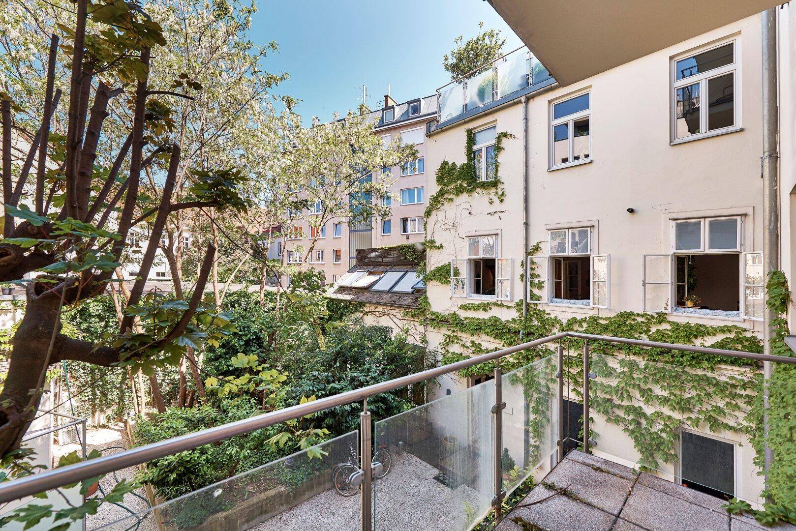 Balkon mit Glasgeländer und Blick auf einen grünen Innenhof und umliegende Gebäude.