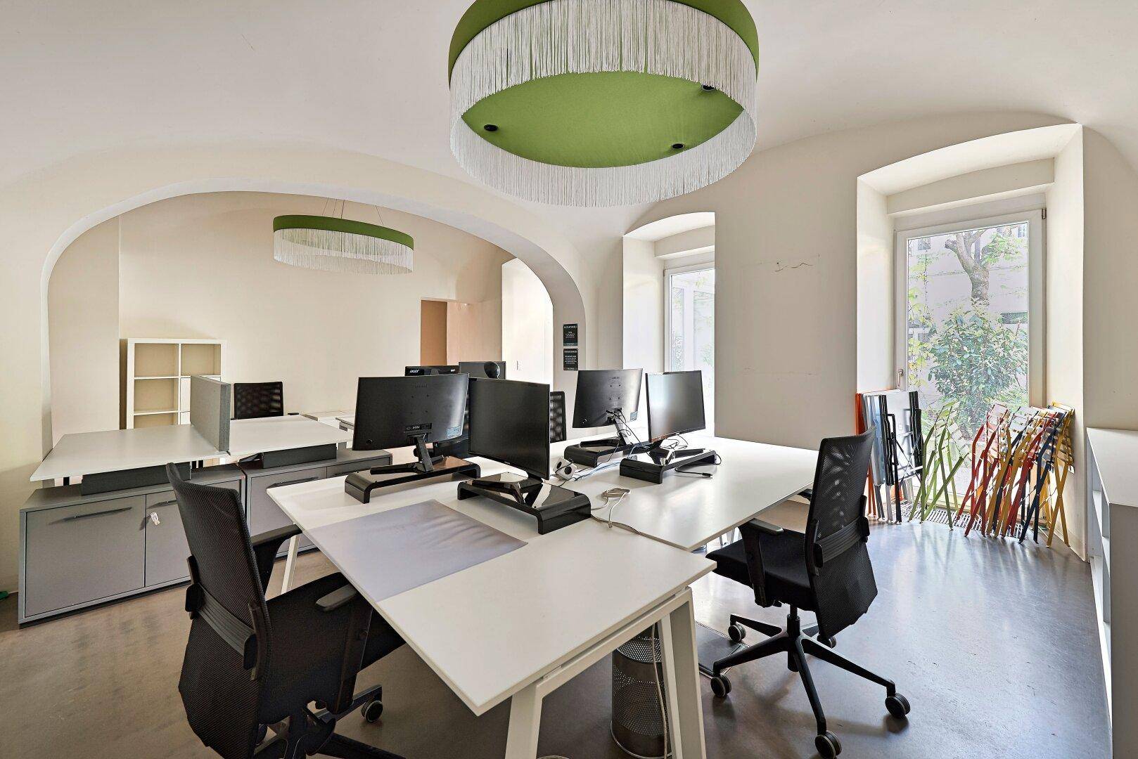 Büroraum mit mehreren Arbeitsplätzen, gewölbten Fenstern und modernen grünen Deckenleuchten.