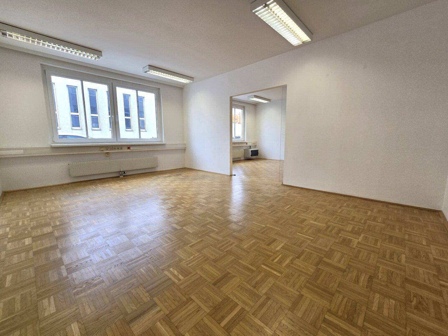 Leeres Büro mit hellem Parkettboden und großen Fenstern, die eine angenehme Arbeitsatmosphäre schaffen.