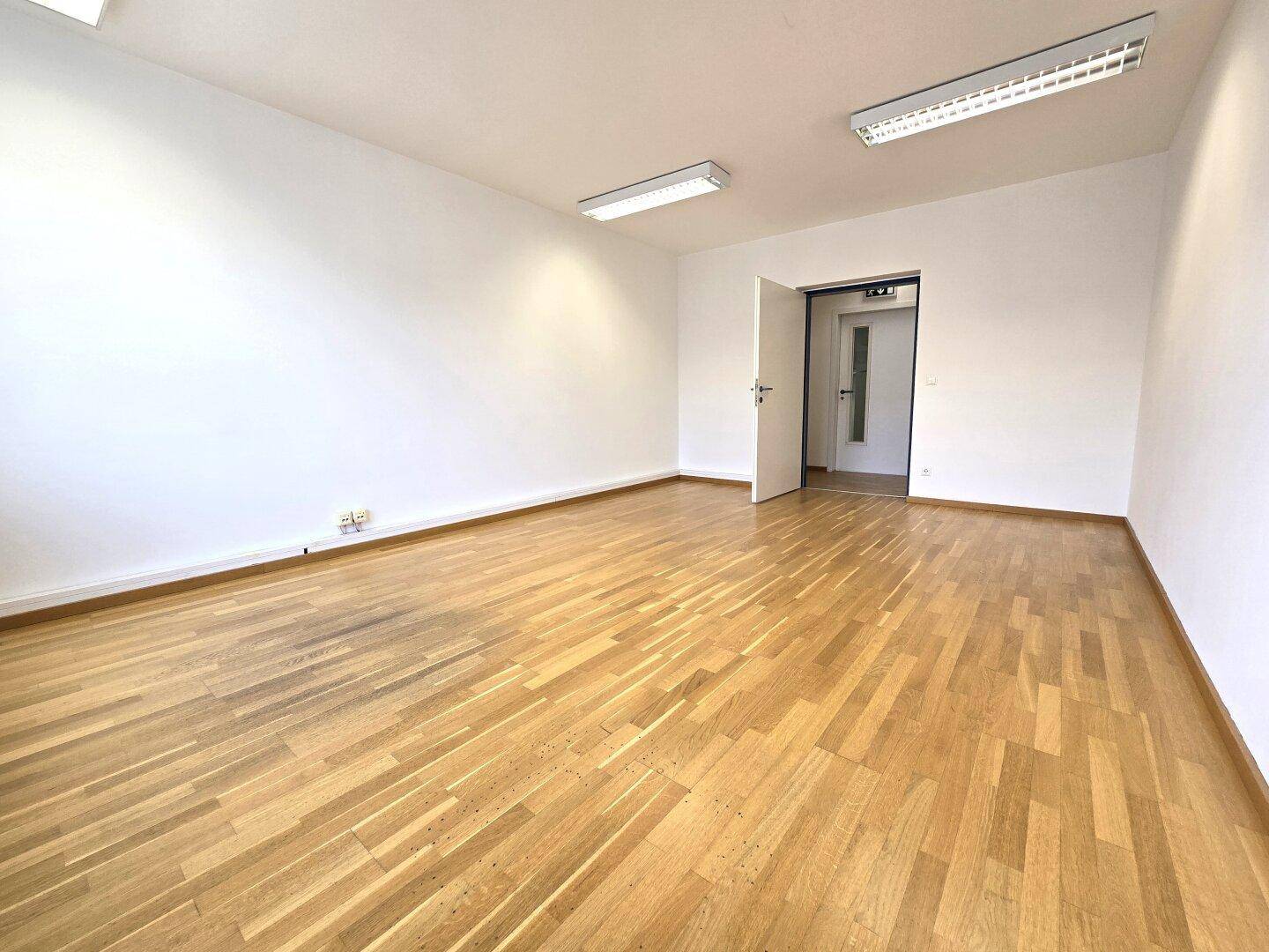 Geräumiger, leerer Büroraum mit Holzboden, weißen Wänden und offener Tür zu einem weiteren Bereich.