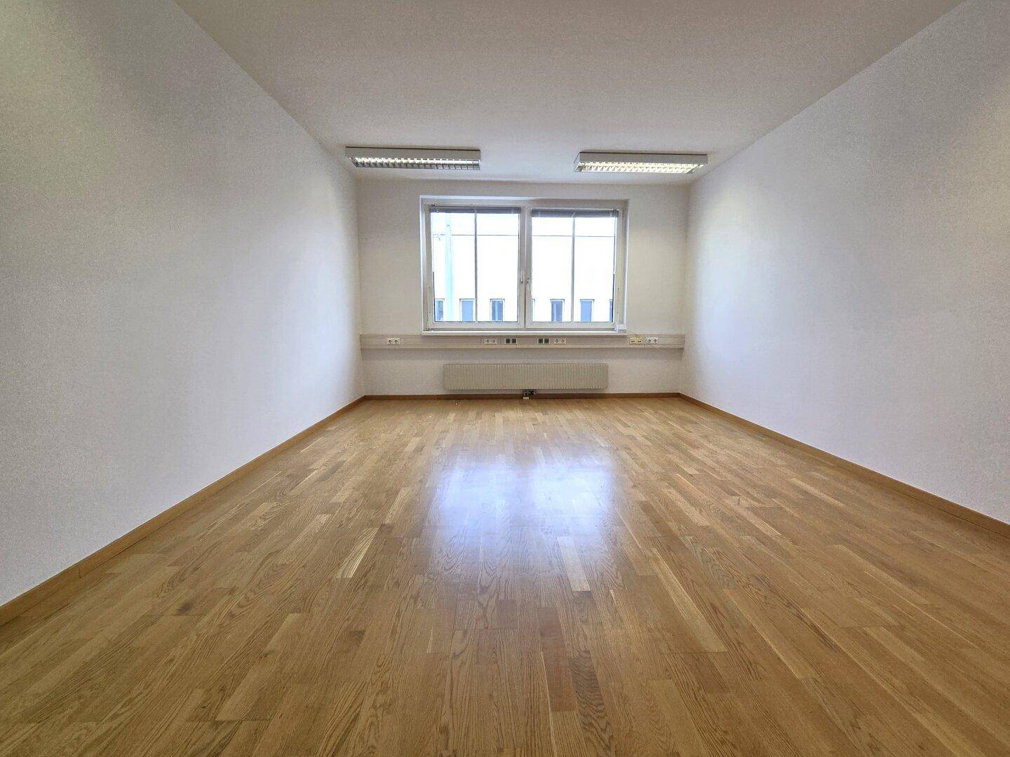 Weitläufiger, leerer Büroraum mit hellem Holzboden, weißen Wänden und einem großen Fenster.