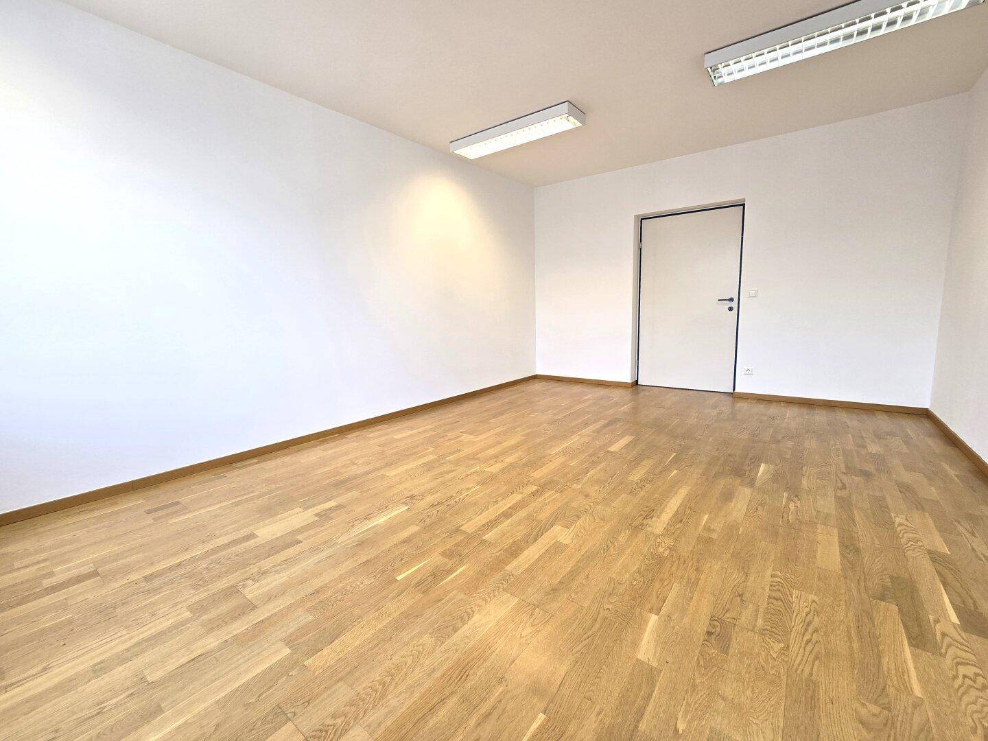 Leerer Büroraum mit Holzboden, weißen Wänden und einer geschlossenen Tür, bereit zur Einrichtung.
