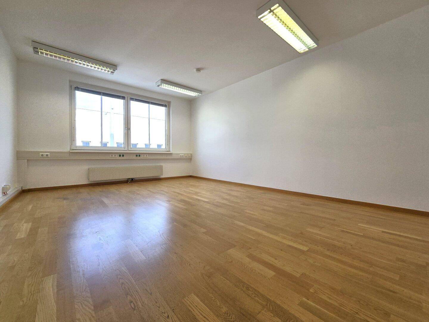 Heller, leerer Büroraum mit glänzendem Holzboden, weißen Wänden und großem Fenster für viel Tageslicht.