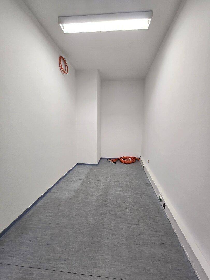 Schmaler, leerer Raum mit grauem Betonboden und weißen Wänden, mit sichtbaren Kabeln an der Seite.