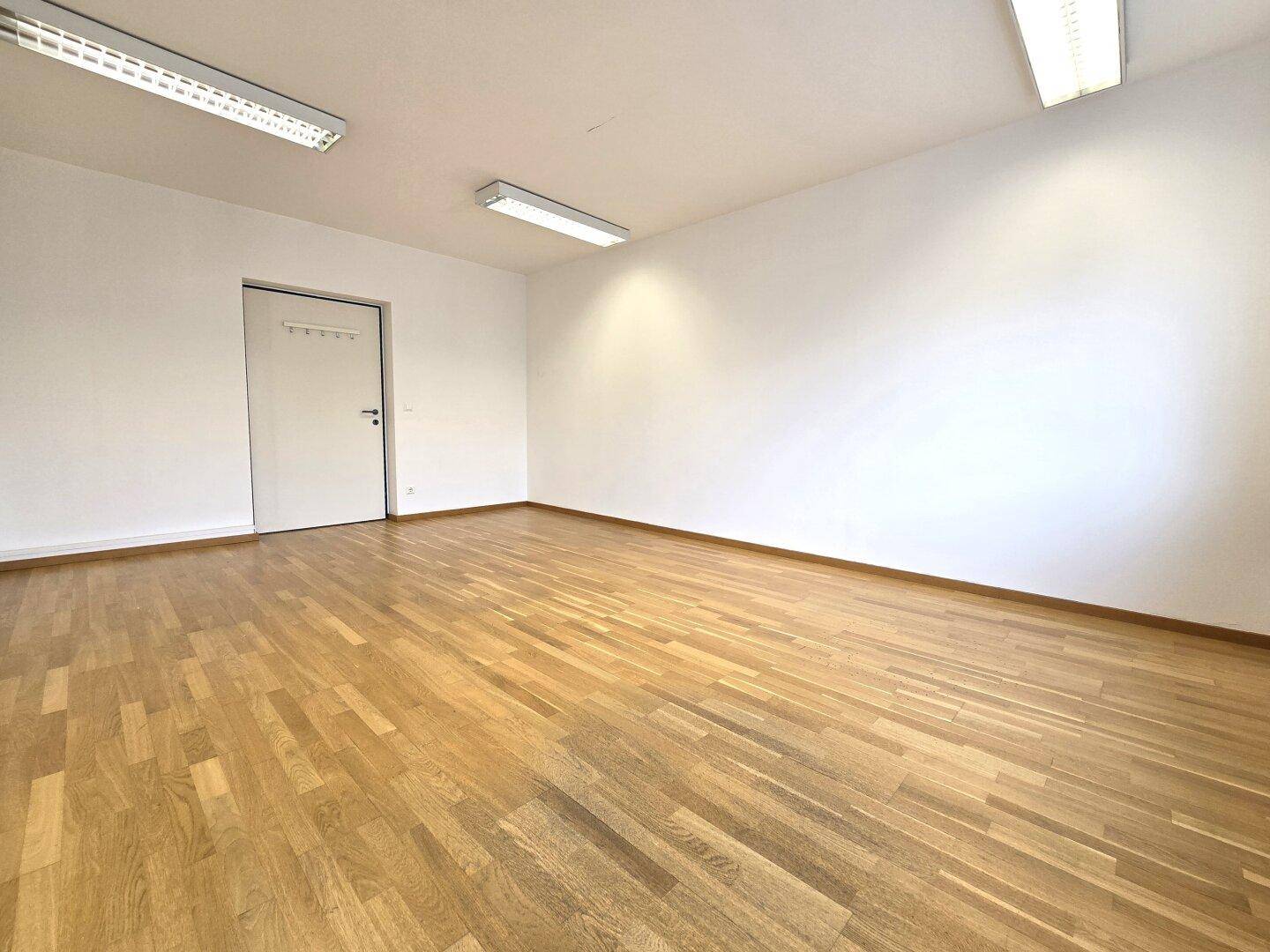 Leerer Büroraum mit Holzboden, weißen Wänden und einer geschlossenen Tür, bereit zur Einrichtung.