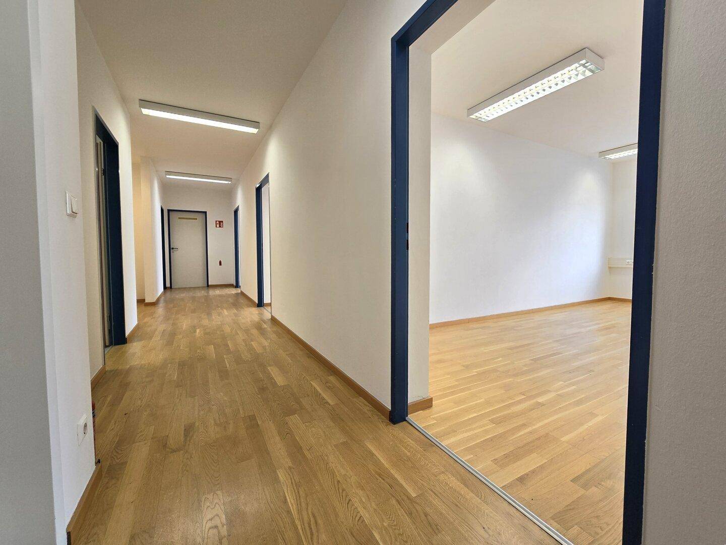 Langer Flur mit Holzboden, weißen Wänden und mehreren Türen, der zu einem hellen Büroraum führt.