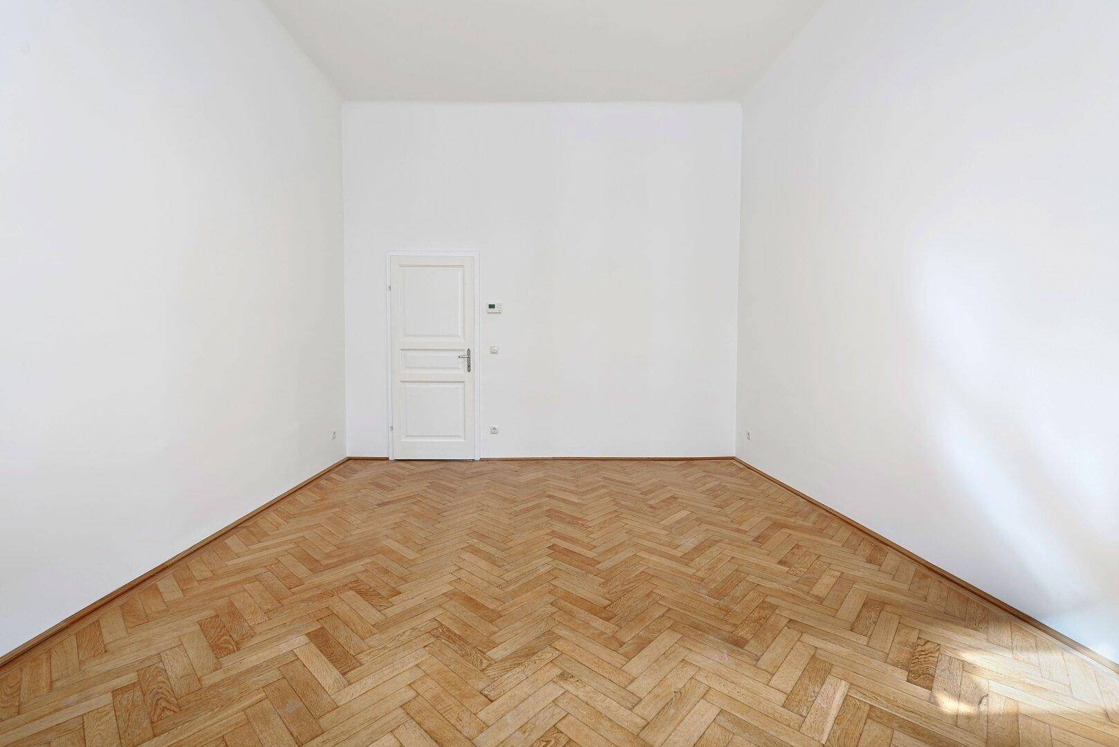 Leeres Zimmer mit hellem Fischgrätparkett und einer weißen Tür, bereit zur Einrichtung.
