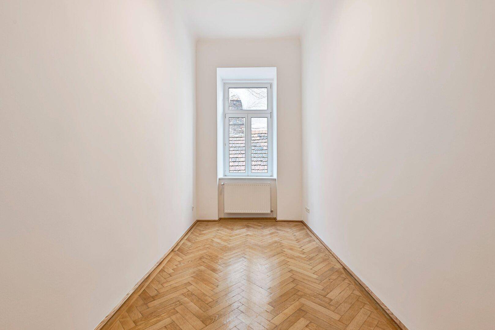 Langes, schmales Zimmer mit Fischgrätparkett und einem Fenster, das Tageslicht spendet.