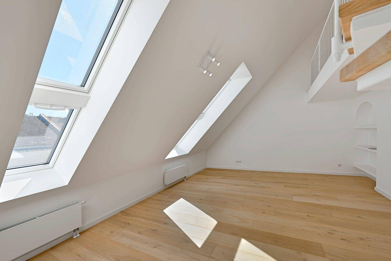 Lichtdurchfluteter Dachgeschossraum mit schrägen Wänden, Holzfußboden und großen Dachfenstern.