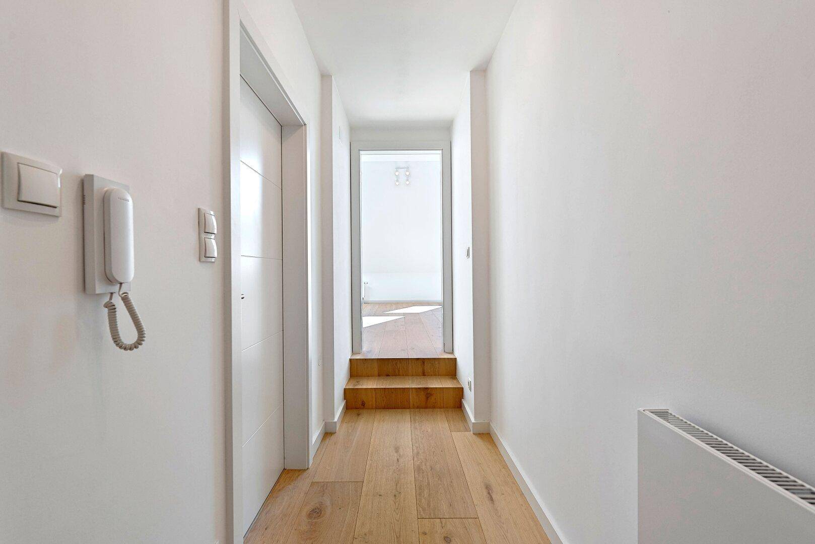 Heller Flur mit Holzfußboden, weißen Wänden und Blick in einen weiteren Raum.