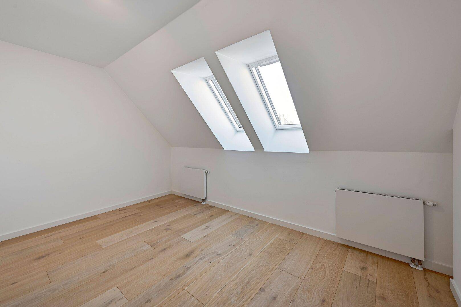Helles Schlafzimmer im Dachgeschoss mit schrägen Wänden, Holzfußboden und zwei Dachfenstern.