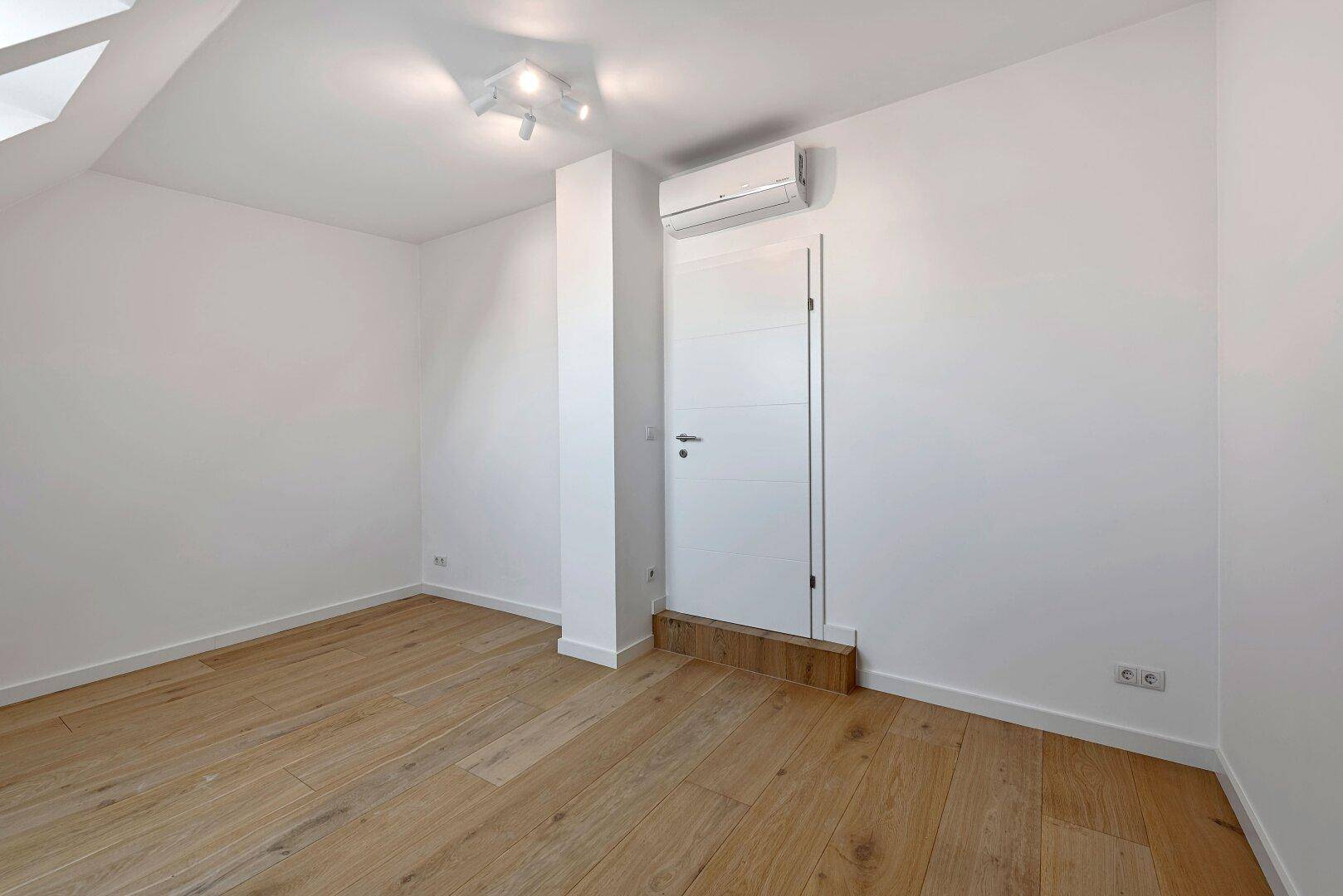 Helles Zimmer mit Holzfußboden, Klimaanlage und weißer Tür.