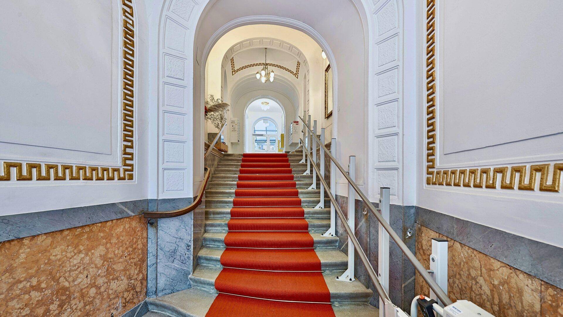 Elegantes Treppenhaus mit rotem Läufer, Marmorverkleidung und kunstvollen Deckenverzierungen.