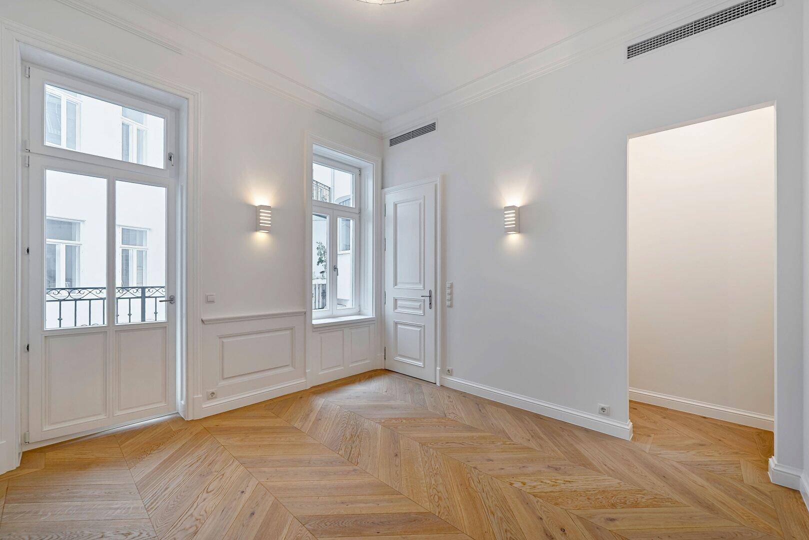 Lichtdurchflutetes Zimmer mit Fischgrätparkett, hohen Fenstern und französischem Balkon.