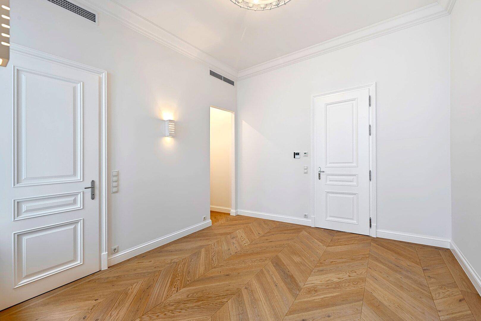 Geräumiges Zimmer mit Fischgrätparkett, hohen Decken und stilvollen weißen Türen.