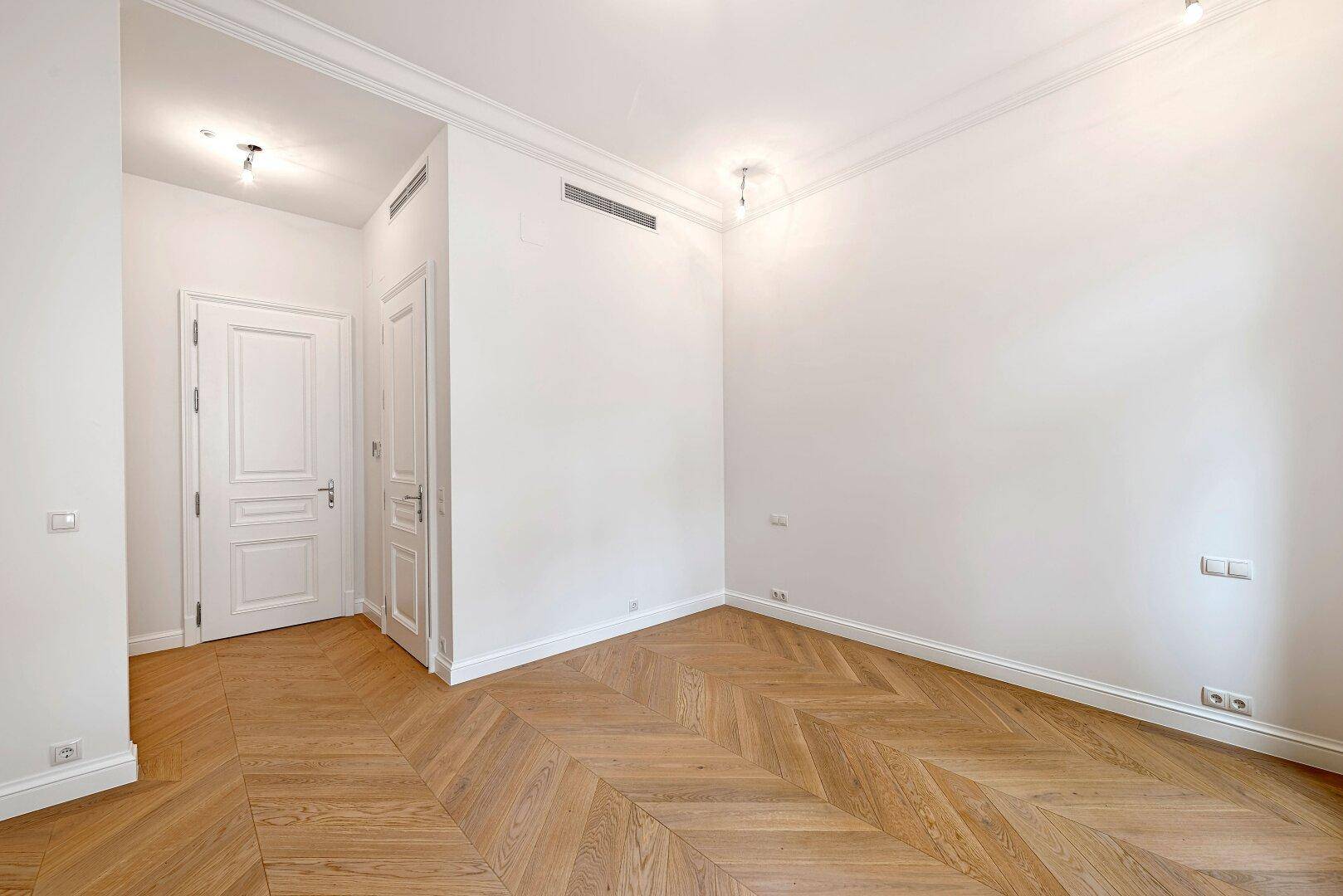 Geräumiges Zimmer mit Fischgrätparkett, hohen Decken und schlichten weißen Wänden.