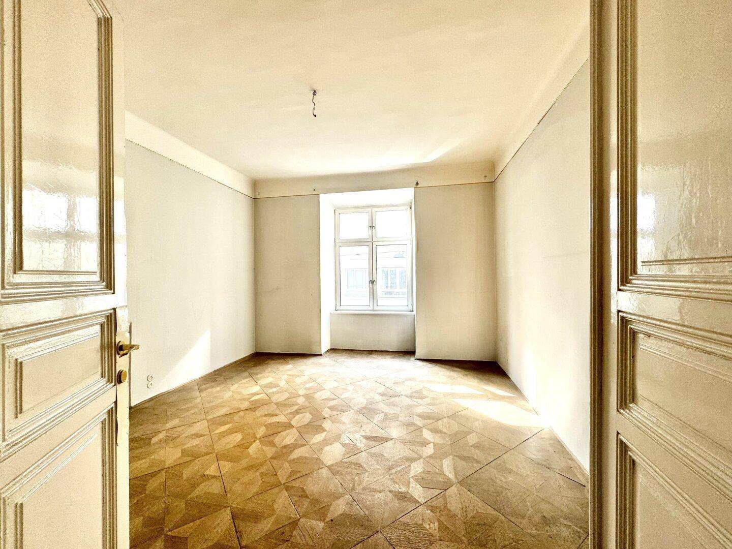 Geräumiges Zimmer mit geometrischem Parkettboden und viel Tageslicht durch das große Fenster.