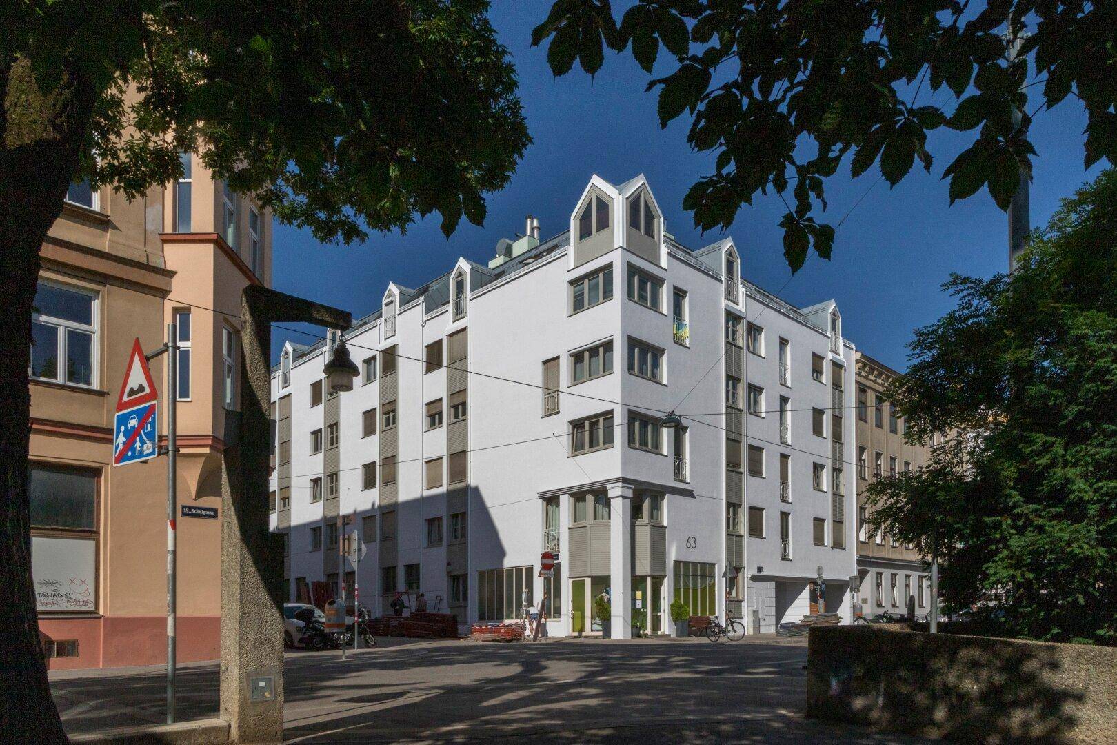 Wohngebäude an einer Straßenecke mit moderner Architektur und vielen Fenstern, umgeben von Bäumen.
