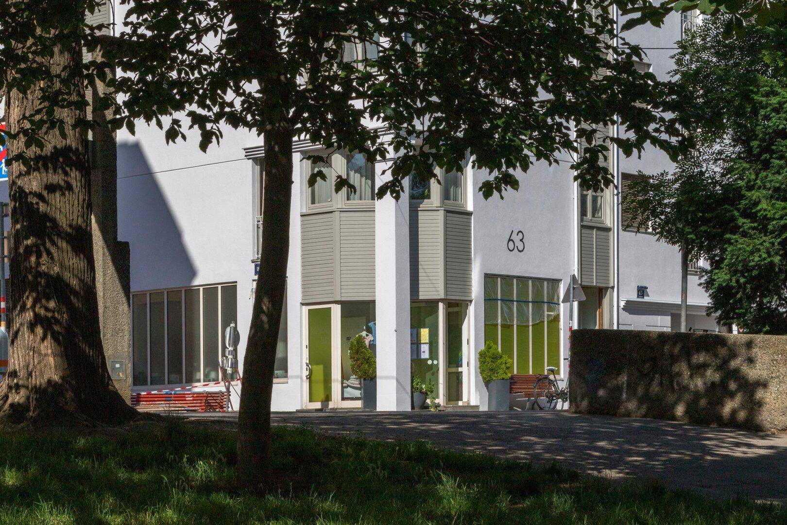 Helle Gebäudefassade mit modernen grauen Erkerfenstern und großen Schaufenstern im Erdgeschoss, umgeben von Bäumen.