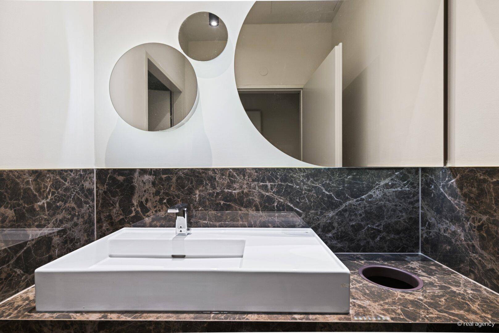 Elegantes Gäste-WC mit einem modernen Waschbecken, einem einzigartigen Spiegeldesign und dunklen Marmoroberflächen.