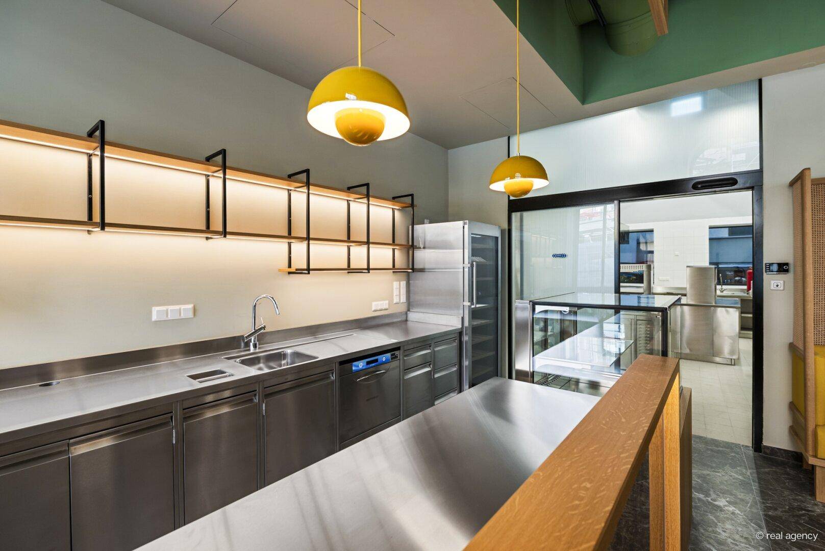 Moderne Küchenzeile mit Edelstahl-Arbeitsflächen, Spüle und Kühlschrank, die durch eine Glastür mit der Hauptküche verbunden ist.