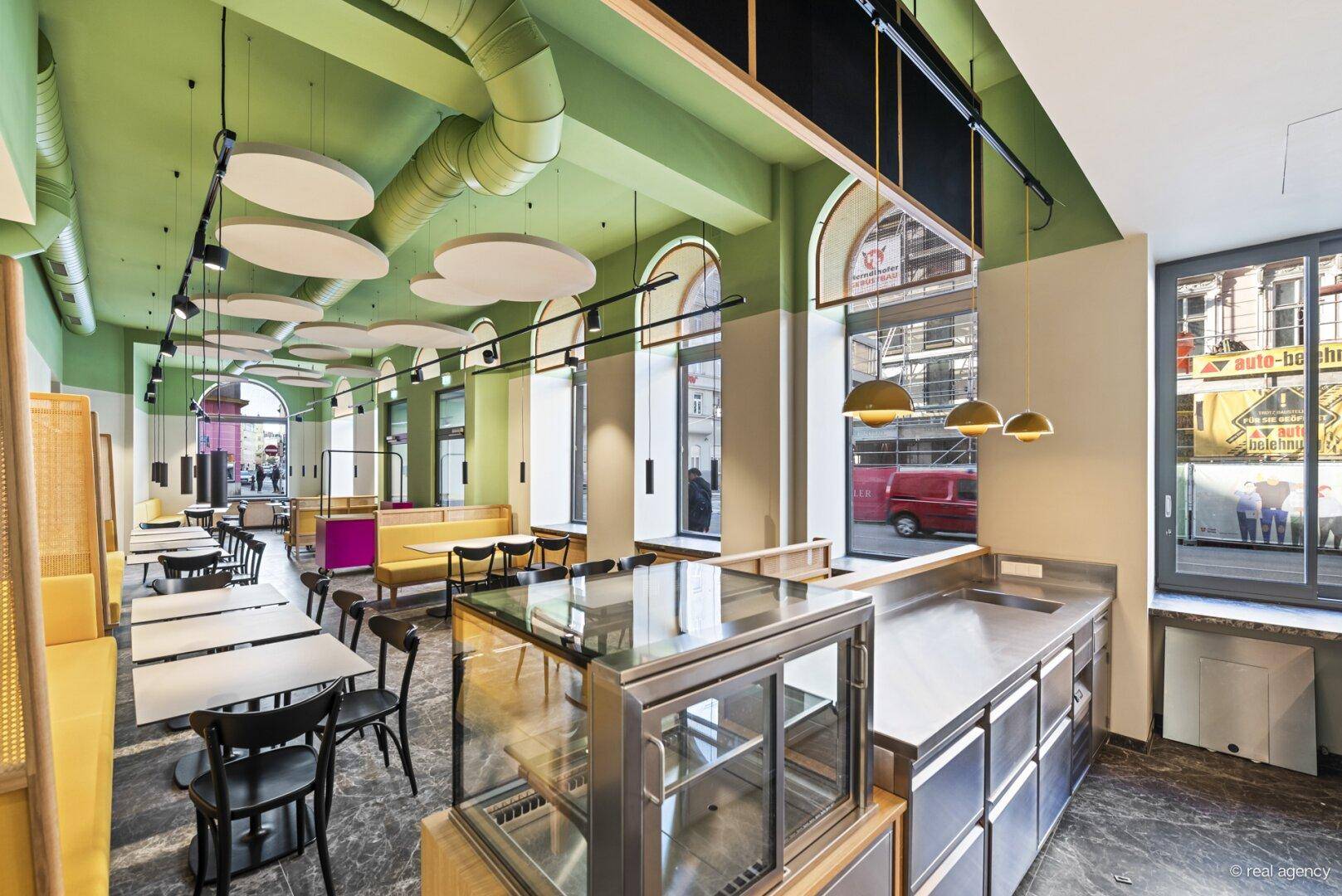 Heller Restaurantbereich mit gelben Sitzbänken, modernen Tischen und einer Theke mit Auslage, die zum Verweilen einlädt.