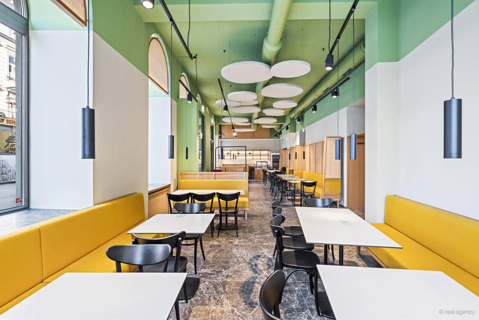 Langer Restaurantbereich mit gelben Sitzbänken, dunklen Tischen und einer grünen Decke, die eine moderne Ästhetik zeigt.