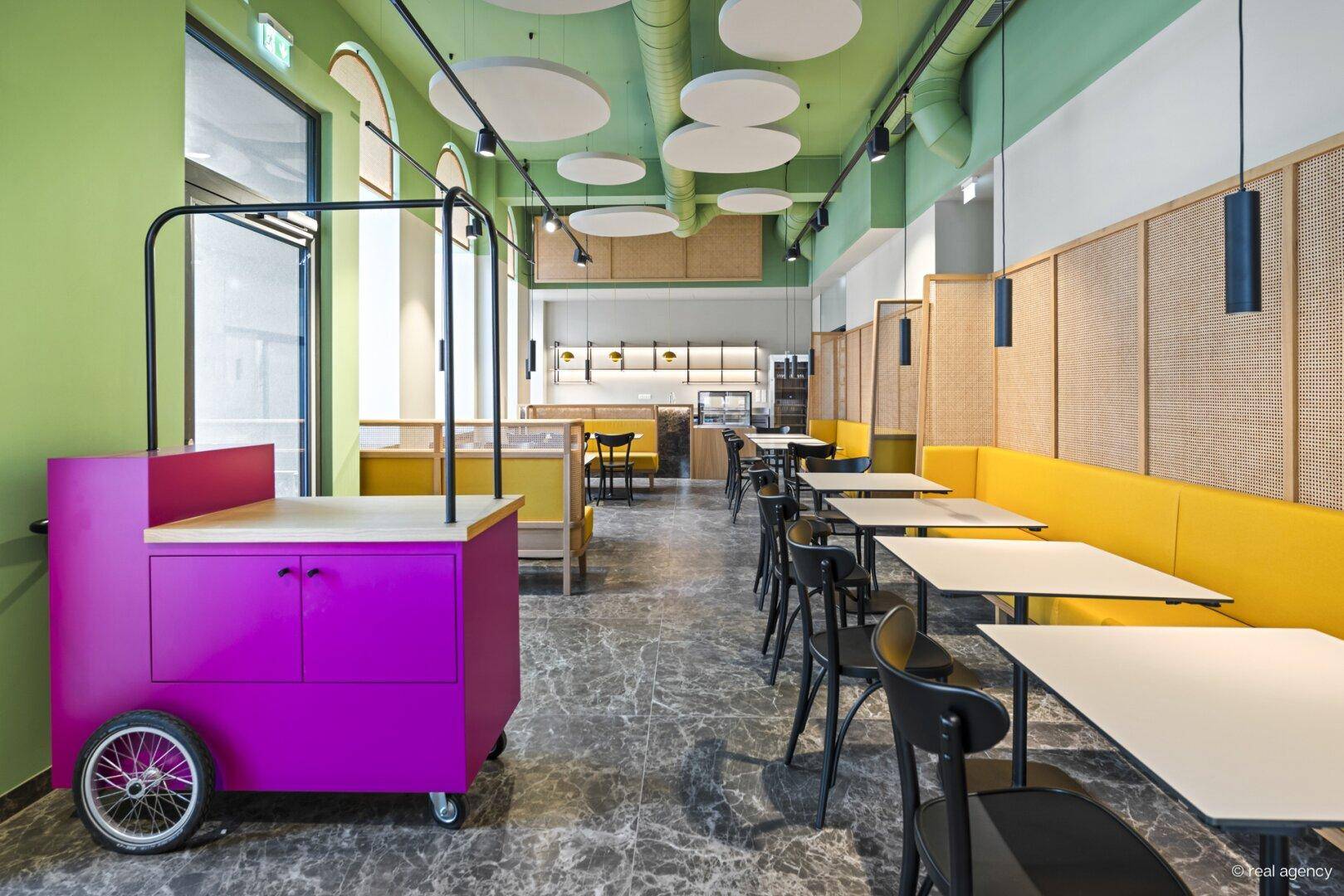 Geräumiger Restaurantbereich mit gelben Sitzbänken, einem mobilen Servierwagen und Blick auf die Theke im Hintergrund.