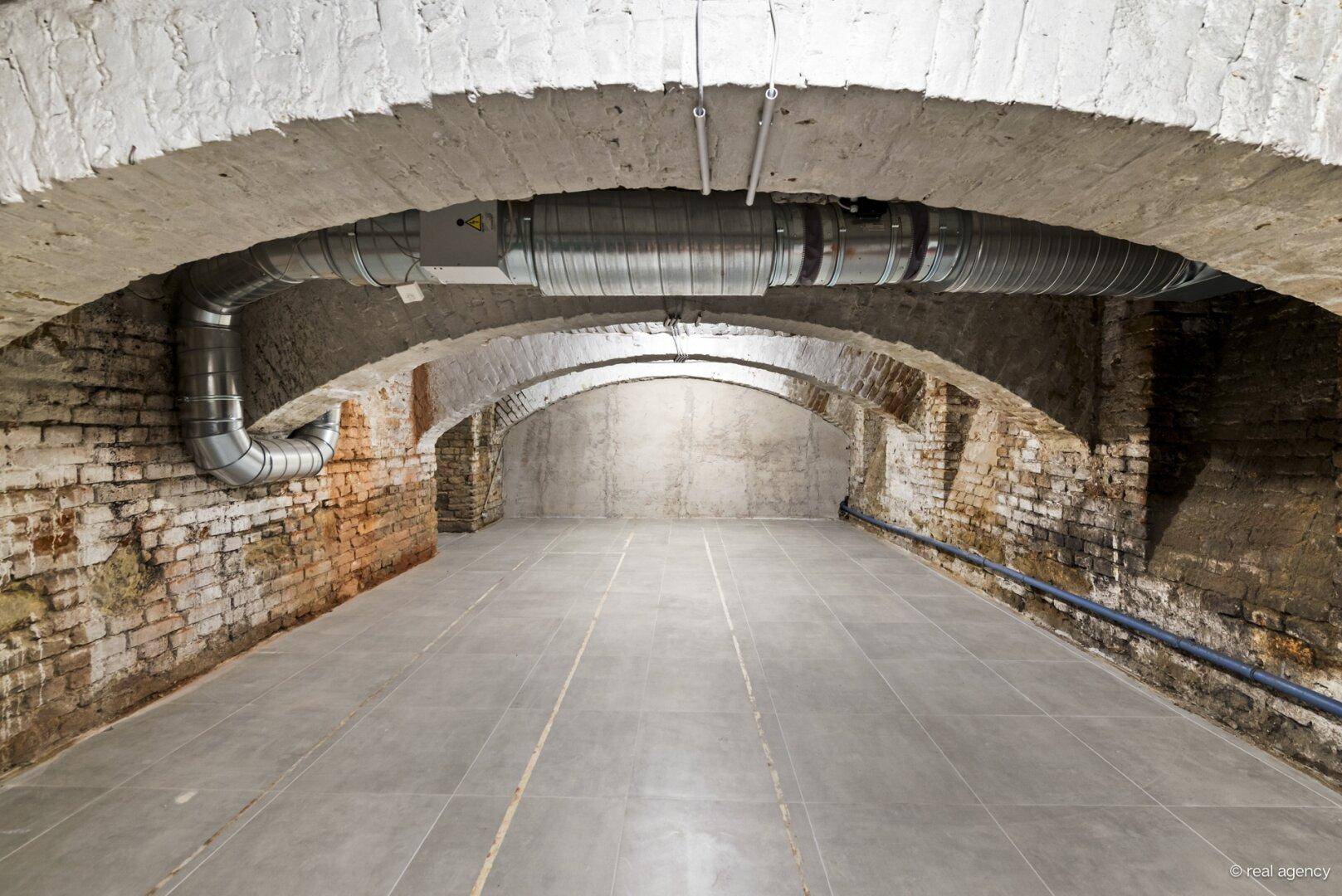 Historischer Gewölbekeller mit unverputzten Ziegelwänden, Fliesenboden und sichtbaren Lüftungsrohren.