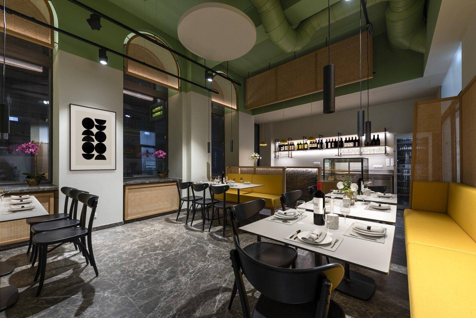 Stilvoller Restaurantbereich mit gelben Sitzbänken, dunklen Tischen und einer Bar im Hintergrund, die eine angenehme Atmosphäre schafft.