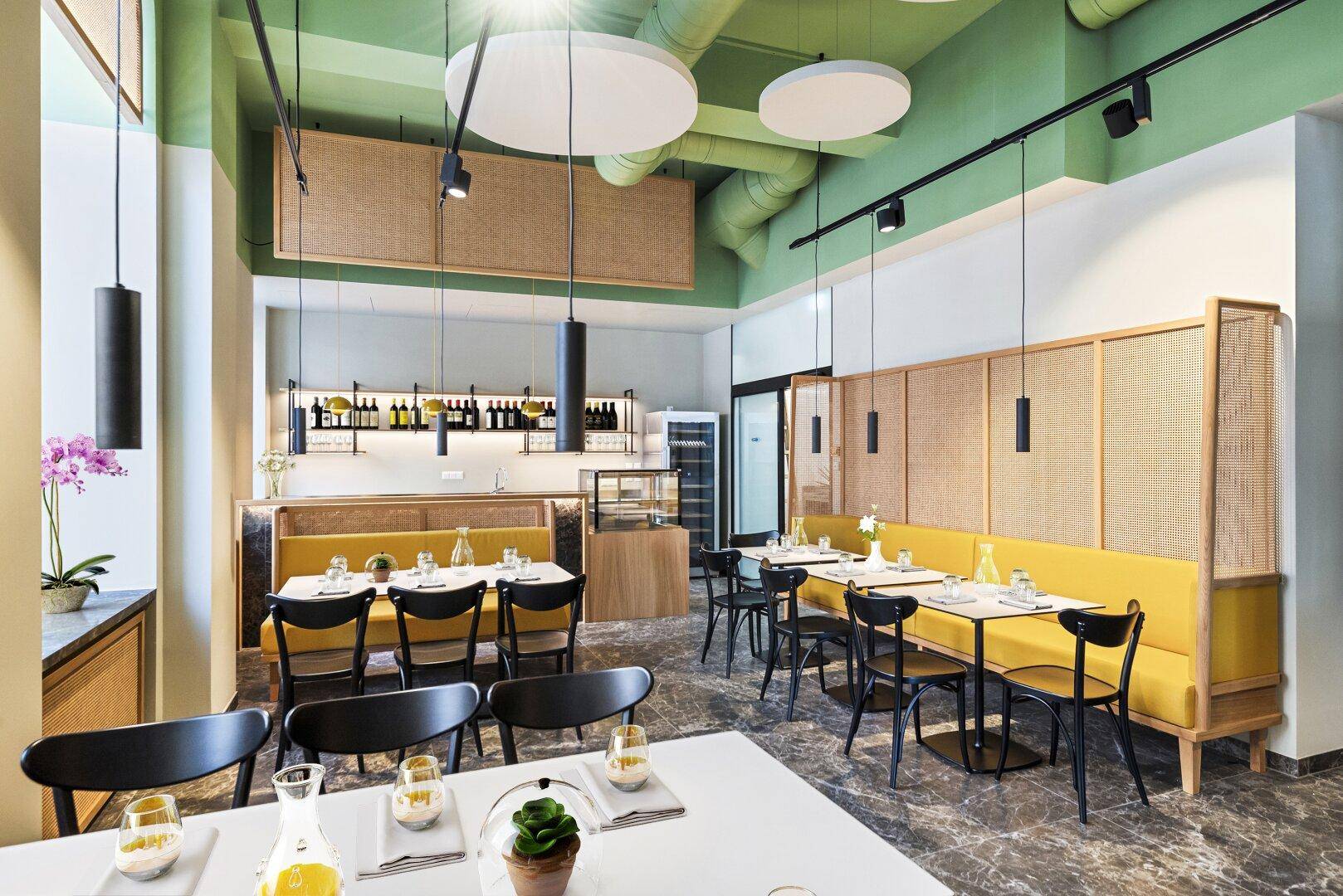 Einladender Restaurantbereich mit gelben Sitzbänken, dunklen Tischen und einer Bar im Hintergrund, die eine gemütliche Atmosphäre schafft.