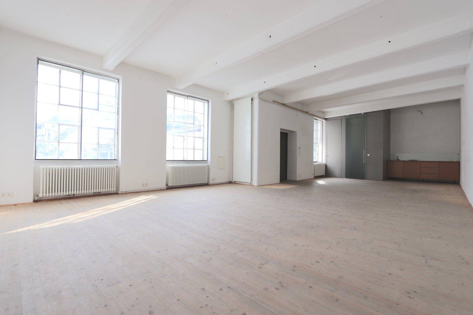 Großer, leerer Raum mit hohen Decken, Holzboden und großen Fenstern, ideal für ein Büro oder Loft.