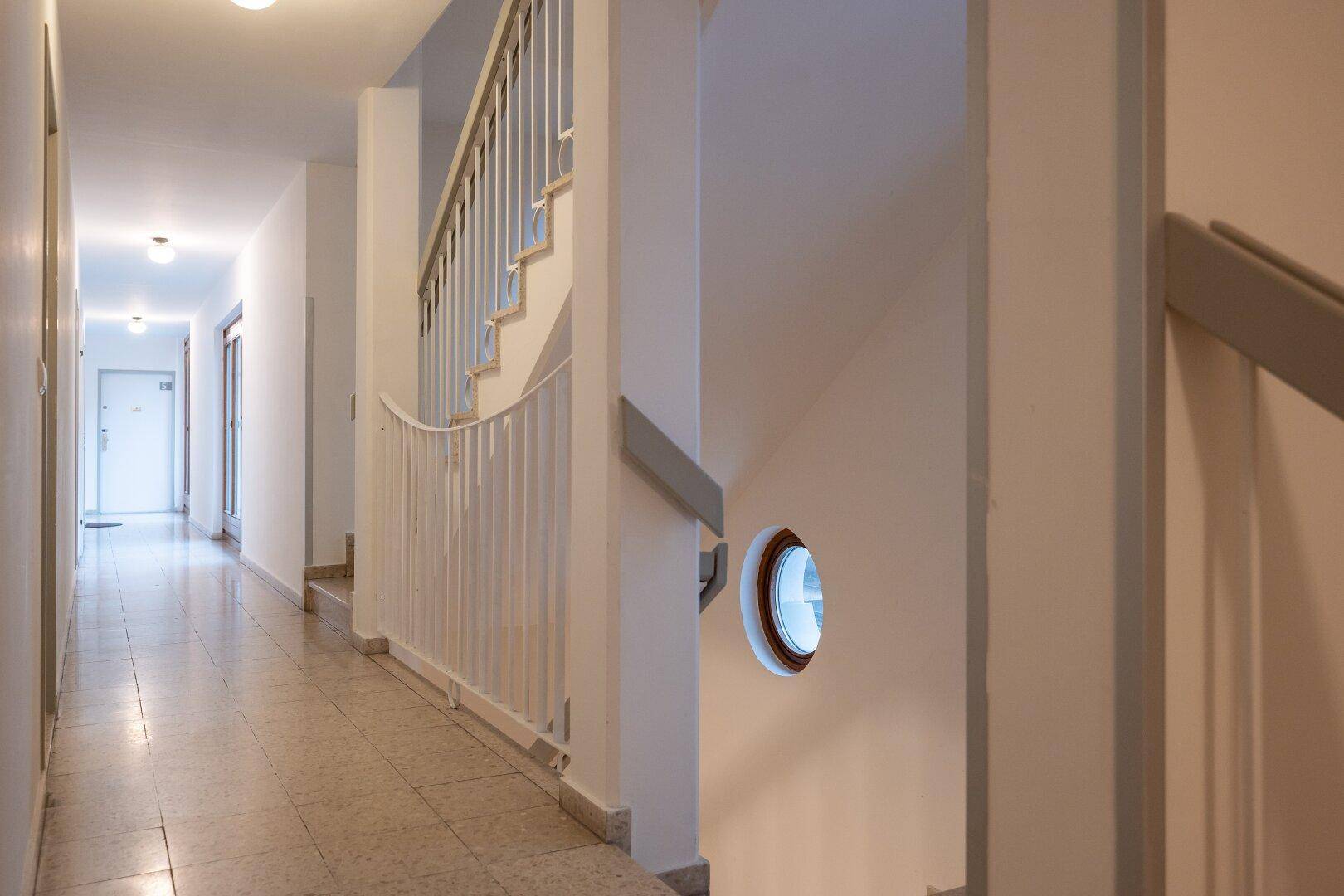Heller Hausflur mit Treppenaufgang, Fliesenboden und einem runden Fenster im Treppenhaus.