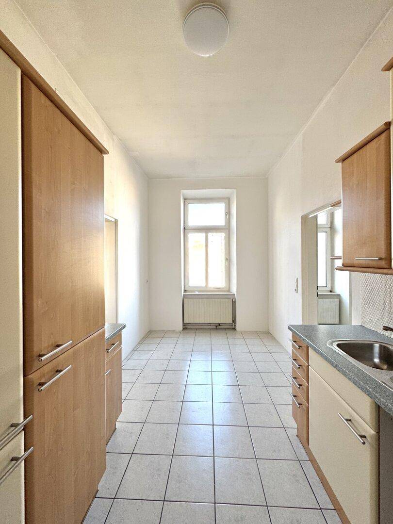 Längliche Küche mit Holzschränken, Fliesenboden und einem Fenster am Ende, das Tageslicht spendet.