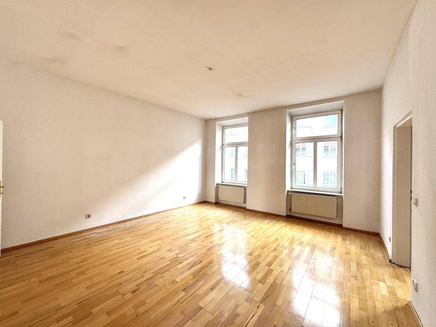 Lichtdurchfluteter Raum mit glänzendem Parkettboden und zwei hohen Fenstern, ideal für Wohn- oder Schlafzimmer.