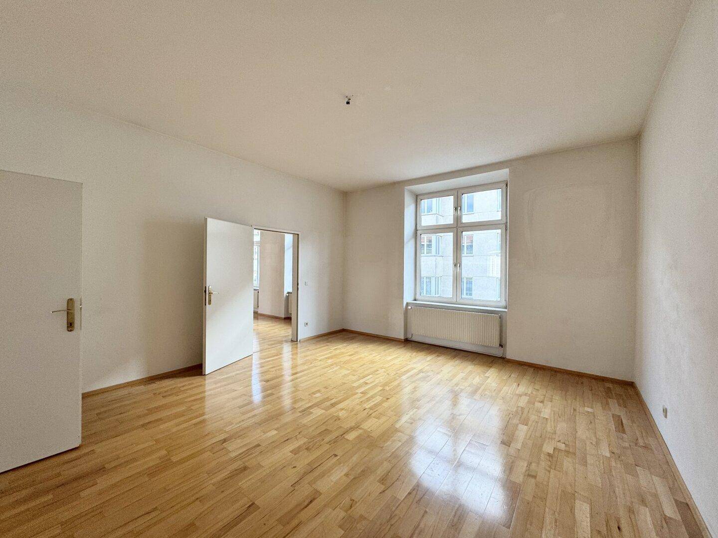 Geräumiges, helles Zimmer mit Parkettboden und einem großen Fenster, das viel Tageslicht hereinlässt.