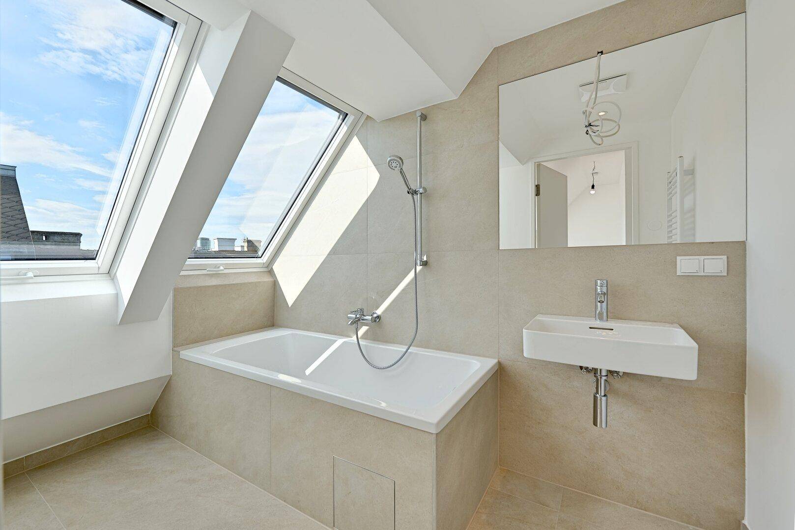 Modernes Badezimmer mit Badewanne, Dachfenstern und beigefarbenen Fliesen, sorgt für helle Atmosphäre.