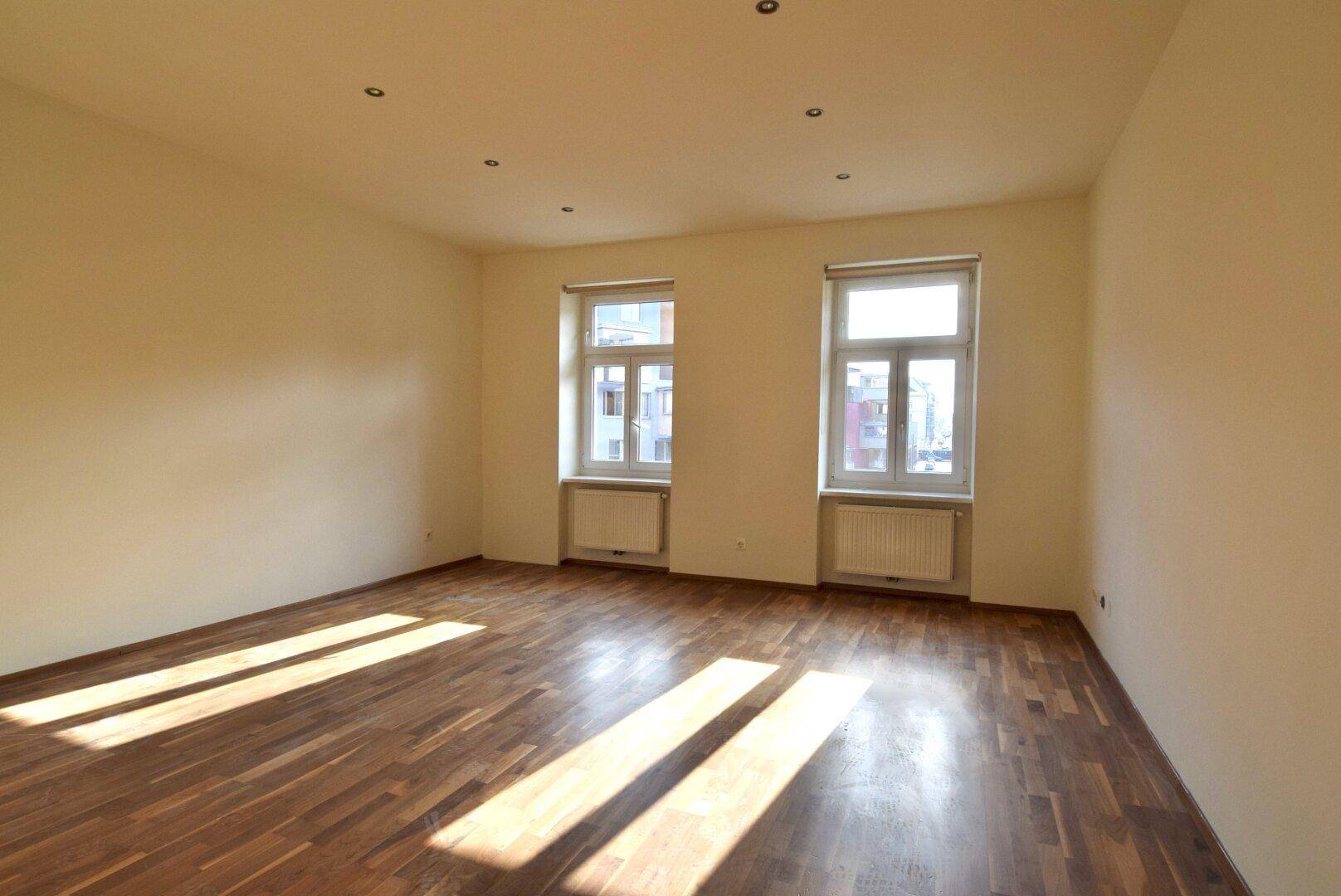 Heller Wohnraum mit warmem Parkettboden und zwei großen Fenstern, die viel Tageslicht hereinlassen.