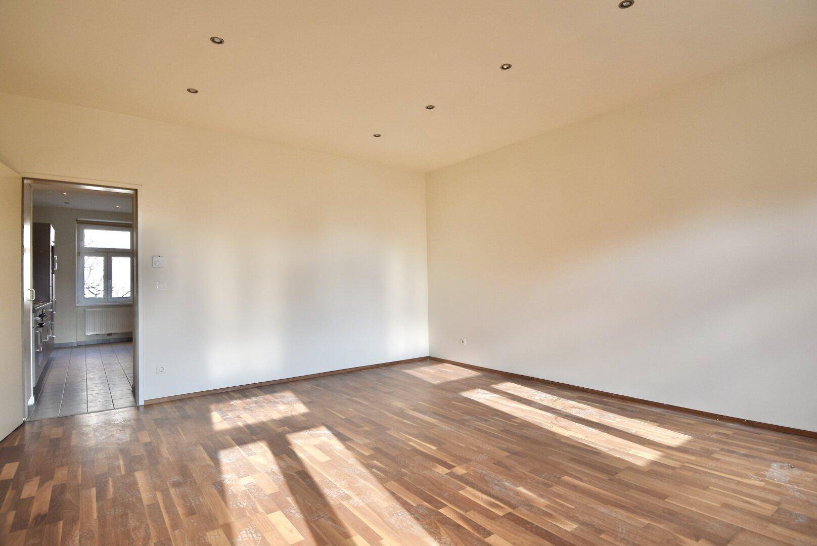 Geräumiger, heller Raum mit Parkettboden und einem Durchgang zu einem weiteren Zimmer, ideal als Wohnbereich.