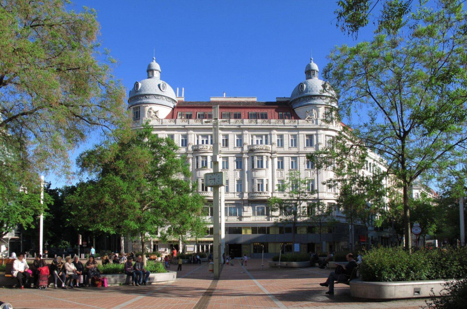 Historisches Gebäude mit Kuppeln und Bäumen auf einem belebten Platz unter blauem Himmel.