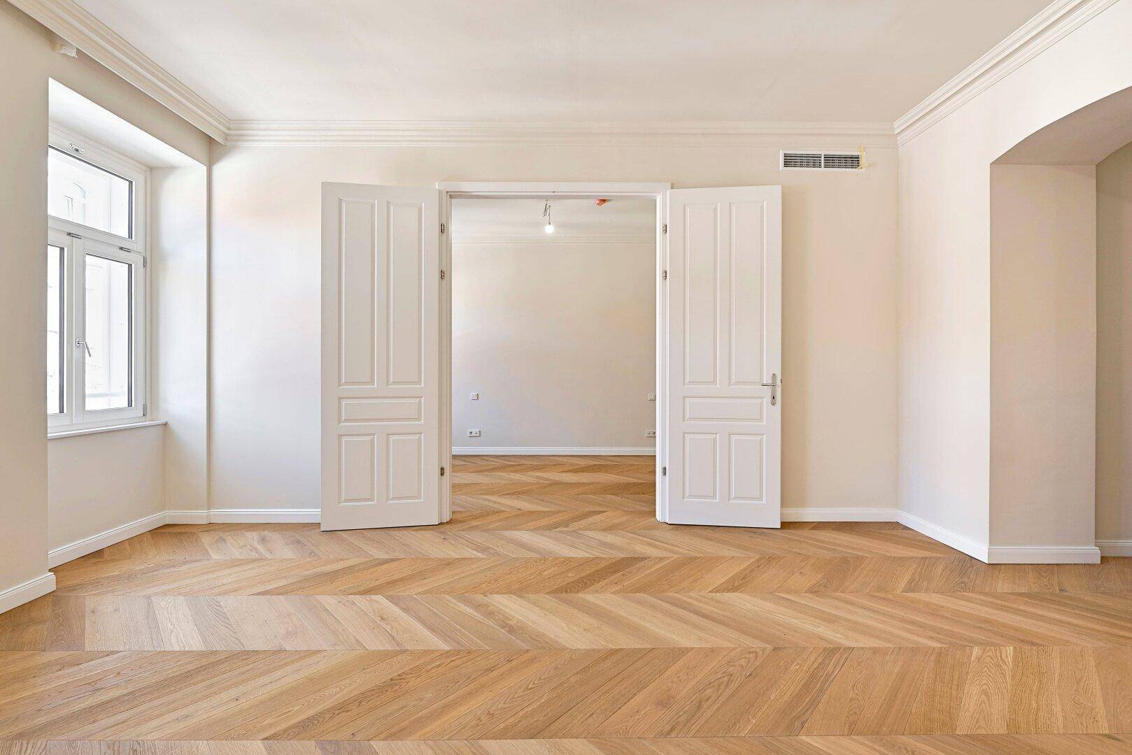 Geräumiges Zimmer mit elegantem Fischgrätparkett und doppelten Flügeltüren zu einem weiteren Raum.