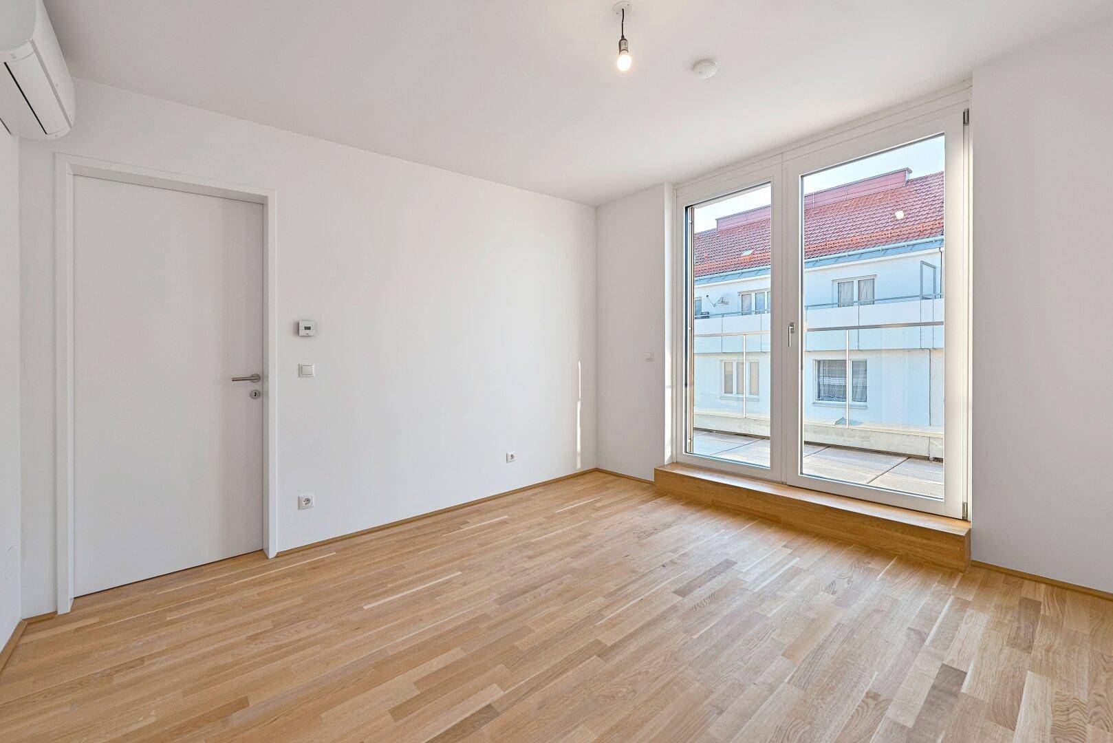 Lichtdurchfluteter Raum mit Holzboden, Klimaanlage und Zugang zu einem Balkon oder einer Terrasse.