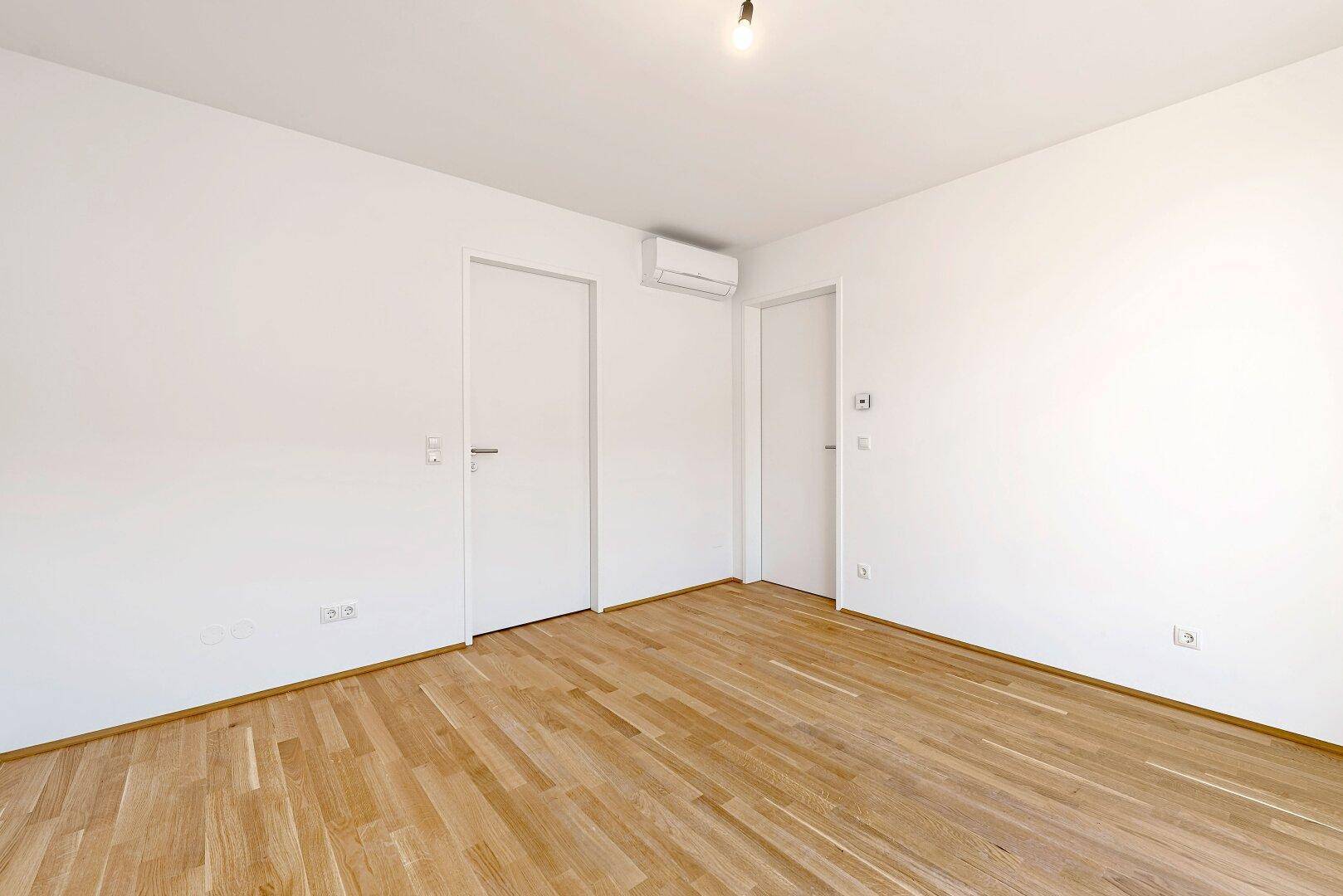 Leerer Raum mit Holzboden, Klimaanlage und mehreren Türen, bereit zur individuellen Nutzung.