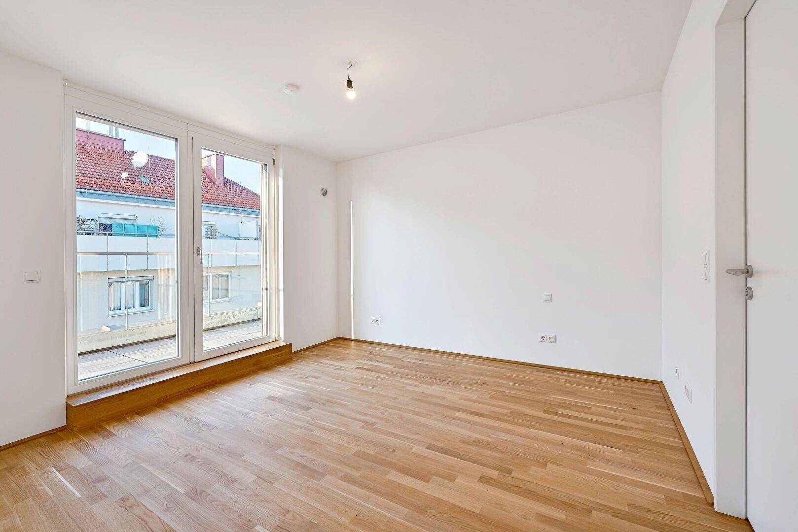 Heller Raum mit Holzboden und bodentiefen Fenstern, die Zugang zu einem Balkon bieten.