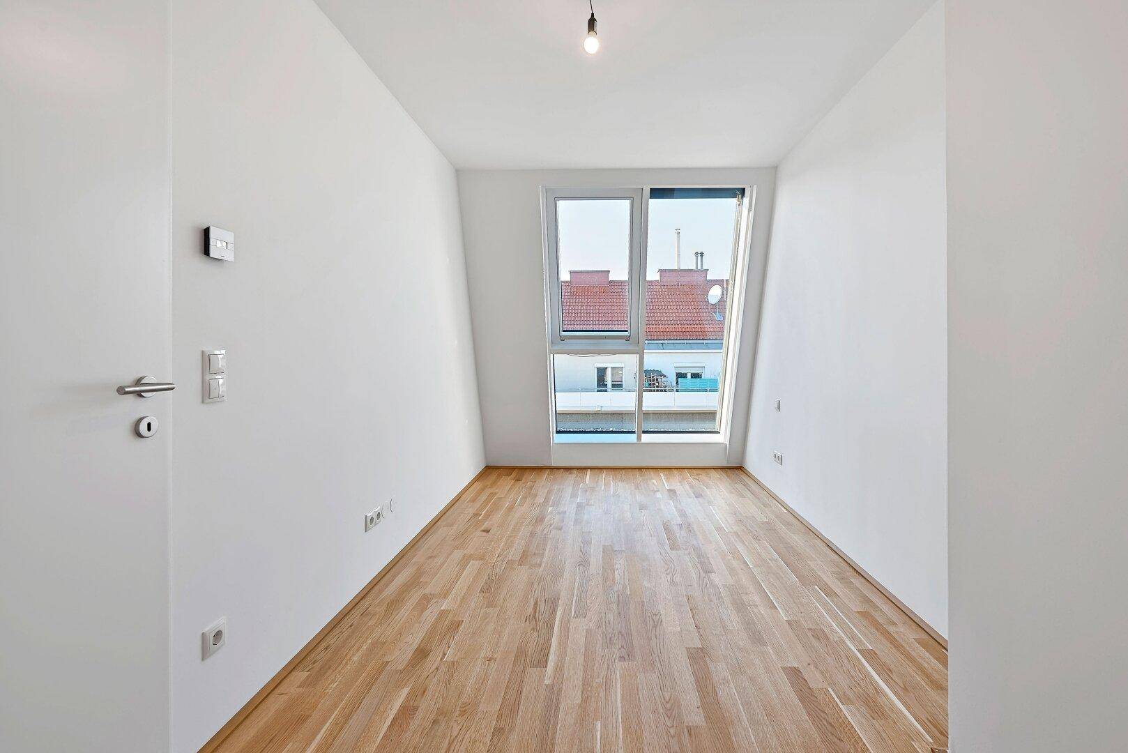 Heller Raum mit Holzboden und großem Fenster, das einen weiten Blick über die Dächer bietet.