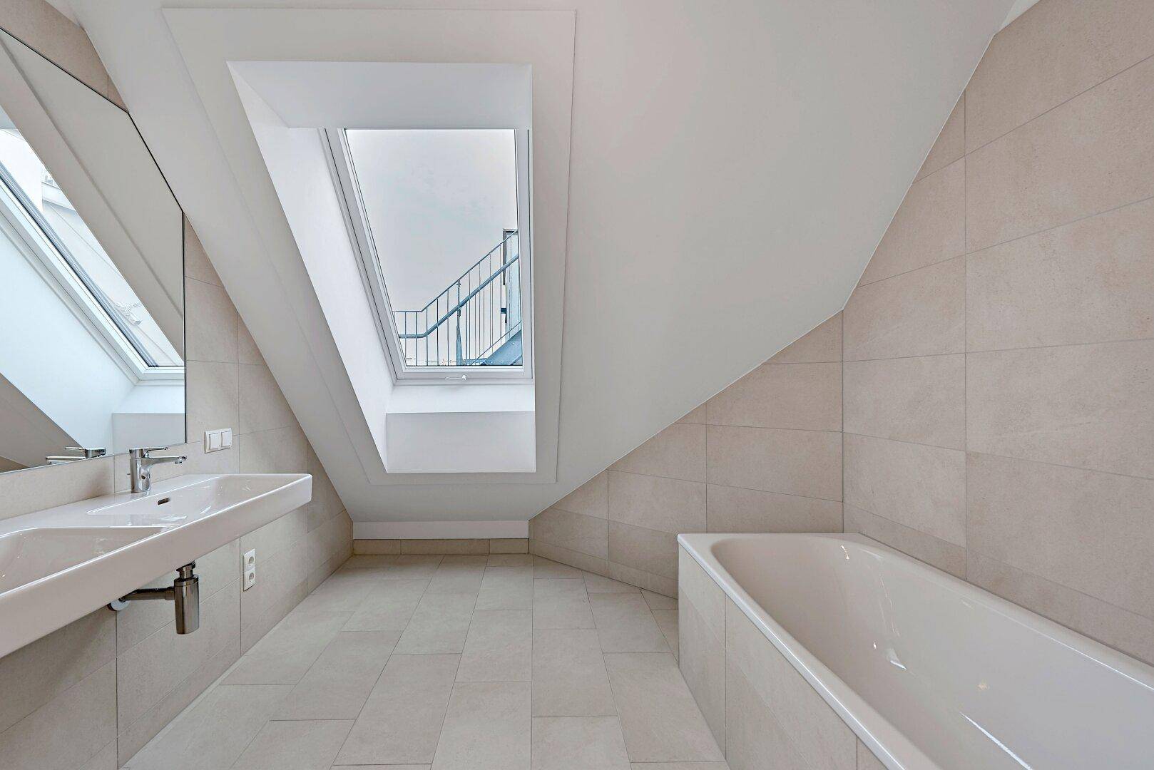 Modernes Badezimmer mit Dachschräge, Dachfenster, Doppelwaschbecken und Badewanne, in hellen Fliesen gehalten.